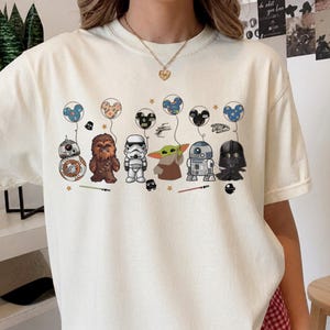 May include: Ivory-colored t-shirt featuring cartoon Star Wars characters holding Mickey Mouse-shaped balloons. Characters include BB-8, Chewbacca, a Stormtrooper, Grogu, R2-D2, and Darth Vader. The design includes stars and lightsabers.