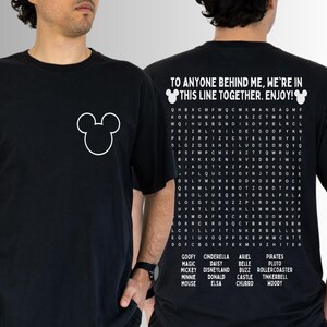 May include: Black t-shirt featuring a white Mickey Mouse outline on the front. The back displays a word search puzzle with Disney-themed words and the phrase "TO ANYONE BEHIND ME, WE'RE IN THIS LINE TOGETHER. ENJOY!"