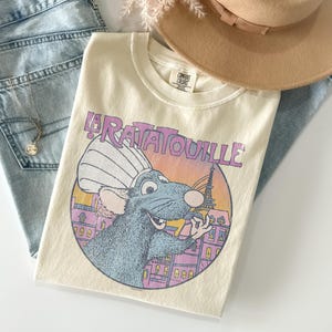May include: Cream-colored t-shirt featuring a cartoon illustration of a rat chef from the movie "Ratatouille" with the Eiffel Tower in the background. The shirt is paired with light-wash denim jeans, a tan hat, and a necklace.