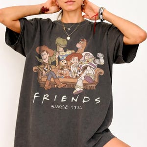 May include: Dark grey t-shirt with a graphic of Toy Story characters on a sofa. The word "FRIENDS" is printed in white, with "SINCE 1995" underneath. The shirt has a vintage, worn appearance.