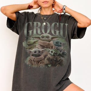 May include: Dark gray t-shirt featuring the word "GROGU" above a graphic of several Grogu characters. The Grogu characters are in various poses and colors, with a soft, faded appearance. The shirt has a vintage, worn look.