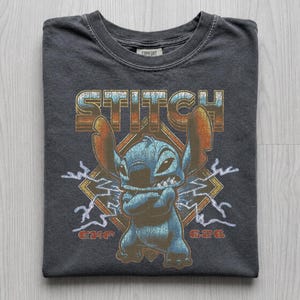 May include: Dark gray t-shirt featuring a graphic of the character Stitch from Lilo & Stitch. The design includes the word "STITCH" in a retro font, Stitch with crossed arms, lightning bolts, and the number "626".