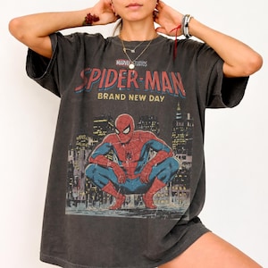May include: A charcoal gray short-sleeve t-shirt featuring a graphic print of the Marvel character Spider-Man in a crouching pose. The text reads Marvel Studios Spider-Man Brand New Day above a city skyline illustration in red, blue, and yellow.