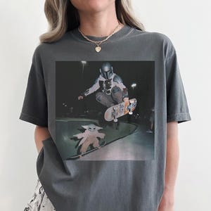 Pu&ograve; includere: Una t-shirt girocollo grigio antracite con una stampa grafica che ritrae un personaggio mandaloriano mentre esegue un trick con lo skateboard in aria sopra una piccola creatura verde su uno skateboard in uno skatepark in cemento di notte.
