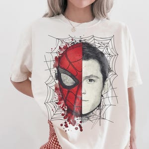 May include: An off-white short-sleeve t-shirt featuring a graphic print of a face split down the middle. One side shows a red and black Spider-Man mask with web patterns and red paint splatters, while the other side shows a realistic human face.