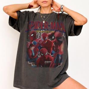 May include: A charcoal gray oversized t-shirt featuring a graphic print of Spider-Man in various poses. The text "SPIDER-MAN" appears in bold red letters at the top, with "BRAND NEW DAY" printed underneath. The design includes lightning bolt accents.