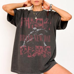 May include: A charcoal grey short-sleeve t-shirt featuring a faded red graphic print of a superhero character in a crouching pose. The design includes multiple smaller character panels and the text BRAND NEW DAY in a bold, distressed red font.