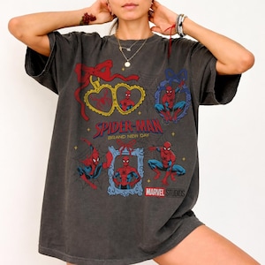 K&ouml;nnte beinhalten: Ein anthrazitfarbenes Oversize-T-Shirt mit mehreren Spider-Man-Grafiken, darunter herzf&ouml;rmige und rechteckige Rahmen mit roten Schleifen, Spinnennetzmustern sowie dem in Rot und Blau gedruckten Text Spider-Man Brand New Day und Marvel Studios.