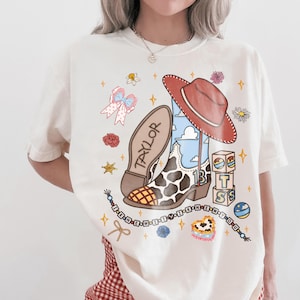 May include: An off-white t-shirt featuring a graphic print of a cowboy boot with a cow print pattern, a red hat, blocks spelling T S, and a beaded string with the text I KNEW IT Y I KNEW YOU. The boot sole is labeled TAYLOR.