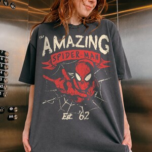 May include: A charcoal gray short-sleeve t-shirt featuring a distressed graphic of Spider-Man in red and white. The text reads The AMAZING SPIDER-MAN and Est. '62, with a spiderweb design integrated into the background of the central character illustration.