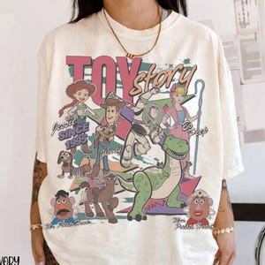 May include: Ivory-colored t-shirt featuring a colorful graphic of Toy Story characters, including Woody, Buzz Lightyear, and Jessie. The text "TOY STORY" is displayed above the characters, with "SINCE 1995" below Jessie.