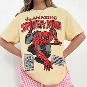 May include: A pale yellow short-sleeve t-shirt featuring a distressed graphic of Spider-Man in a crouching pose. The text reads: the AMAZING SPIDER-MAN, FEATURING PETER PARKER ALIAS SPIDER-MAN, and ANOTHER SPECTACULAR SMASH-HIT FROM THE HOUSE OF IDEAS!
