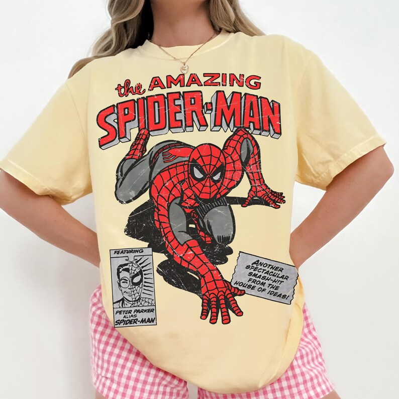 May include: A pale yellow short-sleeve t-shirt featuring a distressed graphic of Spider-Man in a crouching pose. The text reads: the AMAZING SPIDER-MAN, FEATURING PETER PARKER ALIAS SPIDER-MAN, and ANOTHER SPECTACULAR SMASH-HIT FROM THE HOUSE OF IDEAS!