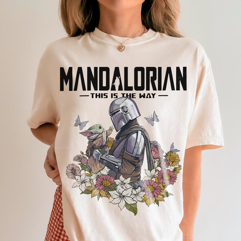 May include: Cream-colored t-shirt featuring the text "MANDALORIAN - THIS IS THE WAY" in black. The graphic depicts the Mandalorian and Grogu surrounded by flowers and butterflies. The design is centered on the shirt.