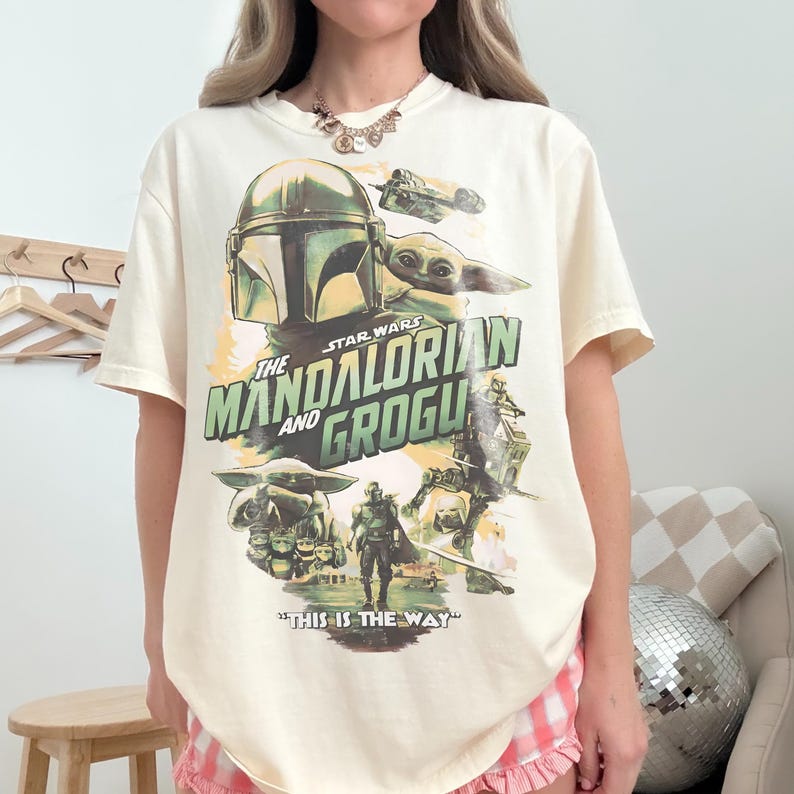 May include: Cream-colored t-shirt featuring a graphic from the Star Wars series, "The Mandalorian and Grogu." The design includes the Mandalorian's helmet, Grogu, and the phrase "THIS IS THE WAY." The shirt has short sleeves and a relaxed fit.