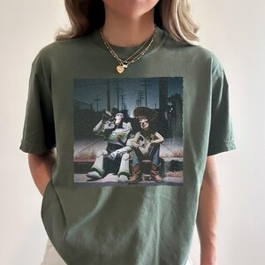 以下が含まれることがあります： オリーブグリーンのTシャツ。トイ・ストーリーのバズ・ライトイヤーとウッディのグラフィックプリント。夜の縁石に座っているキャラクターが描かれています。Tシャツはクルーネックで半袖です。