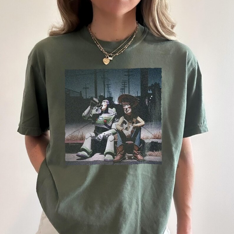 以下が含まれることがあります： オリーブグリーンのTシャツ。トイ・ストーリーのバズ・ライトイヤーとウッディのグラフィックプリント。夜の縁石に座っているキャラクターが描かれています。Tシャツはクルーネックで半袖です。