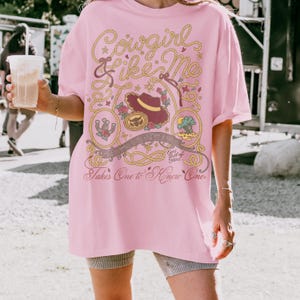 May include: A light pink t-shirt featuring a graphic print with a rope border, a brown cowboy hat, a horseshoe, a desert landscape, and musical notes. The text reads Cowgirl like Me and Takes One to Know One with Eyes full of Stars.