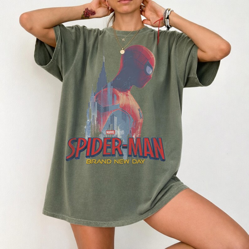 May include: An olive green short-sleeve t-shirt featuring a graphic of Spider-Man in a red and blue suit against a city skyline. The text reads Marvel Studios Spider-Man Brand New Day in red and yellow lettering across the chest area.