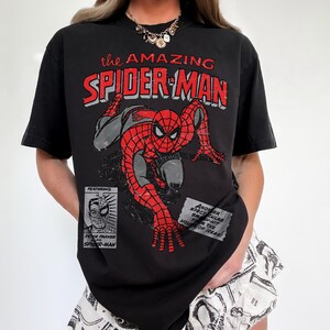 May include: A black short-sleeve t-shirt featuring a distressed graphic of Spider-Man in a crouching pose. The text reads The Amazing Spider-Man, Featuring Peter Parker as Spider-Man, and Another Spectacular Smash-Hit From The House Of Ideas.