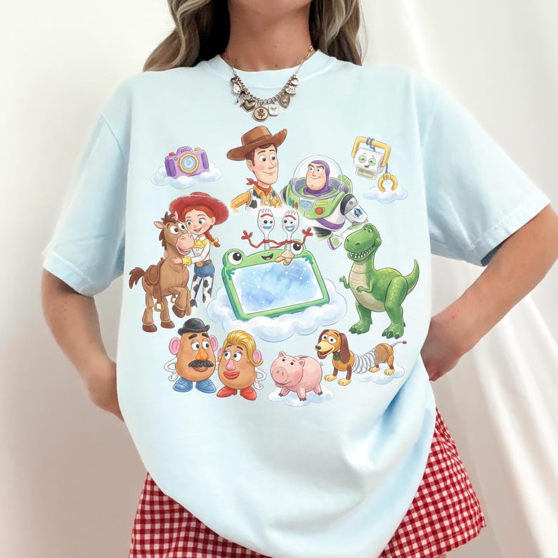 May include: Light blue t-shirt featuring a colorful illustration of characters from the Toy Story movie franchise. The design includes Woody, Buzz Lightyear, Rex, Mr. Potato Head, and others. The shirt is a comfortable, casual style.