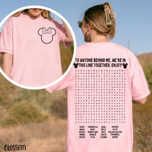 May include: A light pink t-shirt featuring a black Minnie Mouse bow on the front. The back of the shirt has the text "TO ANYONE BEHIND ME, WE'RE IN THIS LINE TOGETHER. ENJOY!" with a word search of Disney characters.