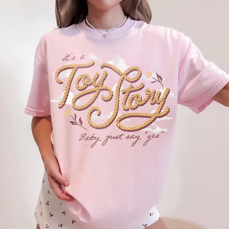 May include: A light pink short-sleeve t-shirt featuring a graphic design with the text "it's a Toy Story" written in a rope-like font, accompanied by the phrase "Baby, just say "yes"" below it, along with illustrations of white clouds and small leaves.