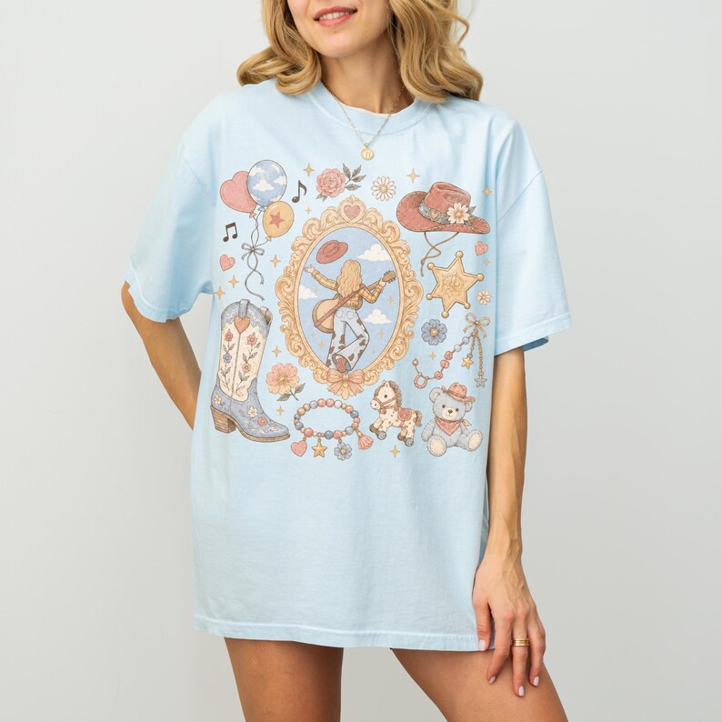 Puede incluir: Una camiseta azul claro con un gr&aacute;fico central de una persona tocando la guitarra dentro de un marco dorado, rodeado de ilustraciones de una bota vaquera, un oso de peluche, un caballito de madera, un sombrero, globos y flores.