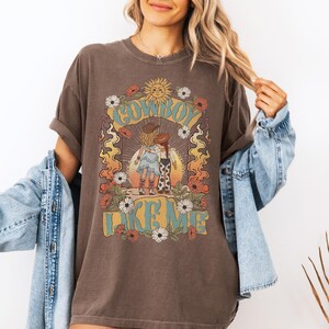 May include: A dark brown short-sleeve t-shirt featuring a graphic print of two figures in cowboy hats and boots standing together. The design includes a sun, flowers, flames, and the text "COWBOY LIKE ME" in a light blue, stylized font.