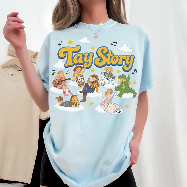 Puede incluir: Una camiseta azul claro con el texto Tay Story en amarillo con borde azul. El gr&aacute;fico incluye varios personajes ilustrados con diversos atuendos, sentados sobre nubes blancas, rodeados de estrellas amarillas y notas musicales negras.