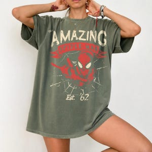 May include: An olive green short-sleeve t-shirt featuring a distressed graphic print of Spider-Man in red with a white spiderweb background. The text reads The AMAZING SPIDER-MAN and Est. '62 in a vintage-style font across the chest area.