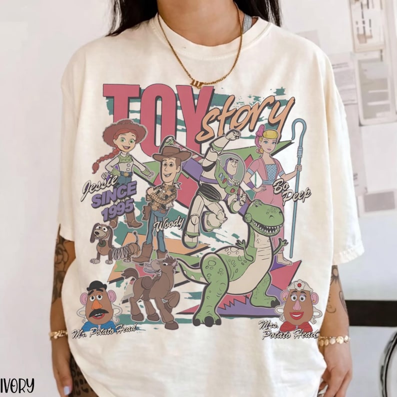 May include: Ivory-colored t-shirt featuring a colorful graphic of Toy Story characters, including Woody, Buzz Lightyear, and Jessie. The text "TOY STORY" is displayed above the characters, with "SINCE 1995" below Jessie.