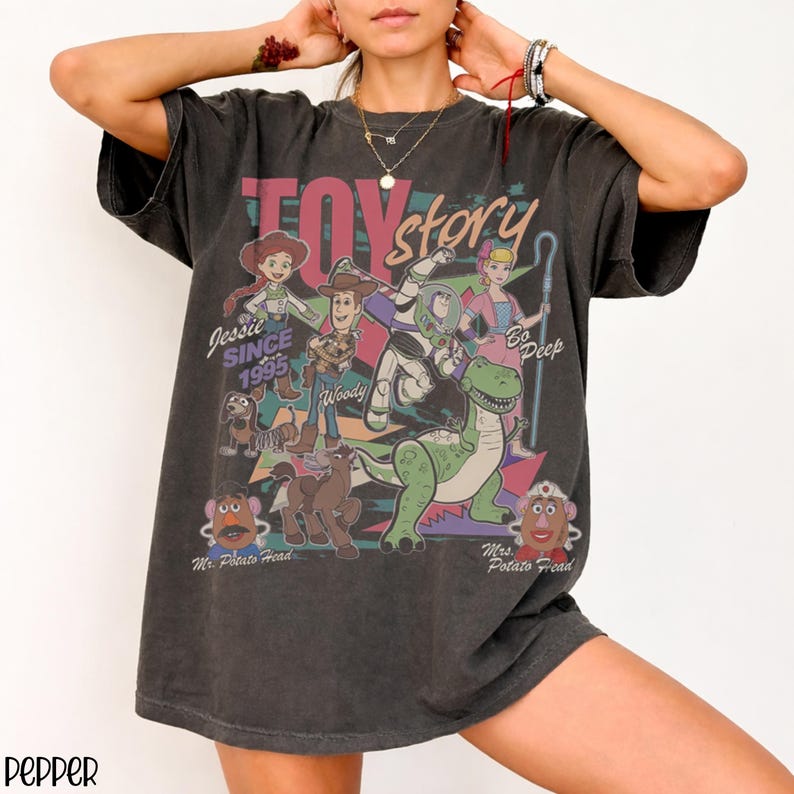 May include: Dark gray t-shirt featuring a colorful graphic of Toy Story characters, including Woody, Buzz Lightyear, and Jessie. The text "TOY STORY" is across the top, with "Jessie Since 1995" and "Go Deep" also visible. The shirt has a vintage look.