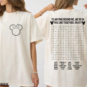 May include: Ivory-colored t-shirt featuring a black Minnie Mouse bow graphic on the front. The back of the shirt has the text "TO ANYONE BEHIND ME, WE'RE IN THIS LINE TOGETHER. ENJOY!" and a word search with Disney characters.