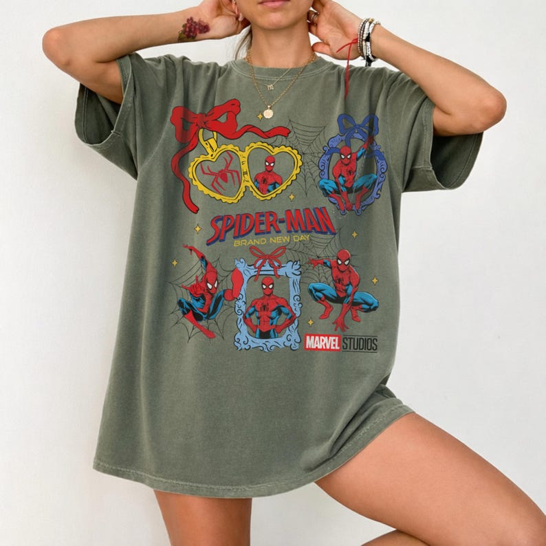 K&ouml;nnte beinhalten: Ein olivgr&uuml;nes Kurzarm-T-Shirt mit einem Grafikdruck von Spider-Man in verschiedenen Posen, eingerahmt von Herz- und Ornamentformen mit roten Schleifen. Der Text lautet Spider-Man Brand New Day und Marvel Studios, erg&auml;nzt durch Spinnennetz-Motive.
