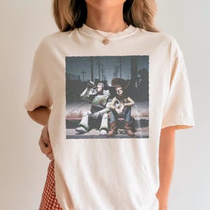 以下が含まれることがあります： クリーム色のTシャツ。トイ・ストーリーのバズ・ライトイヤーとウッディの、かすれたグラフィックが特徴です。キャラクターは縁石に座っており、バズはボトルから飲んでいます。画像はヴィンテージで、少しざらついた雰囲気があり、ノスタルジーを醸し出しています。