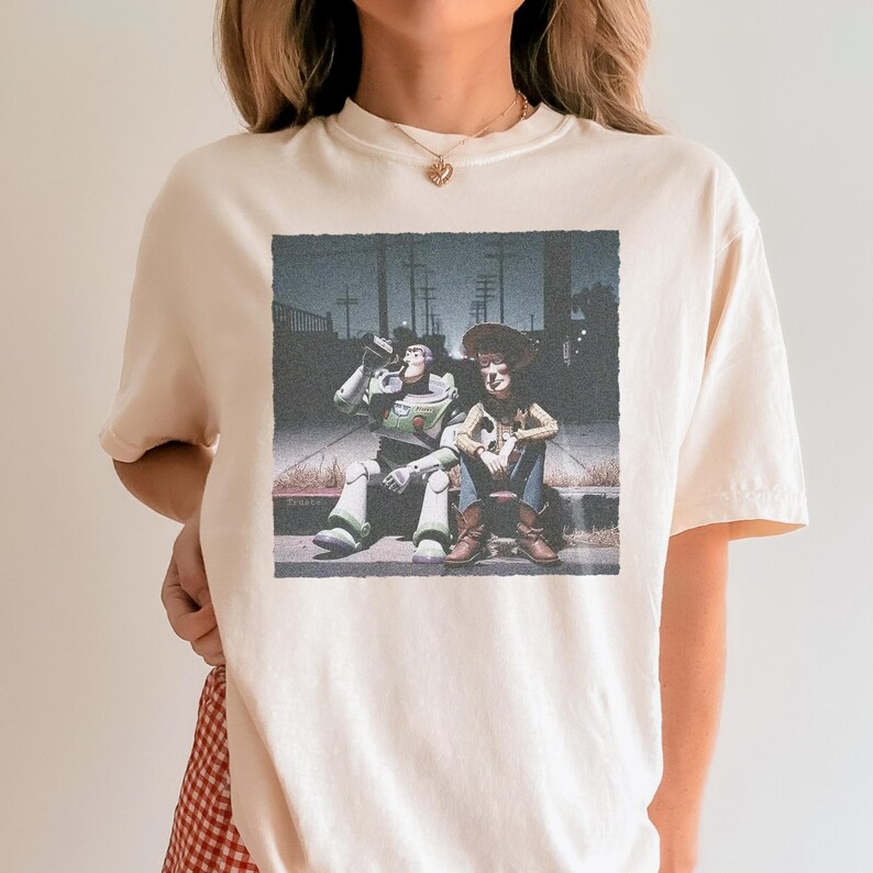 以下が含まれることがあります： クリーム色のTシャツ。トイ・ストーリーのバズ・ライトイヤーとウッディの、かすれたグラフィックが特徴です。キャラクターは縁石に座っており、バズはボトルから飲んでいます。画像はヴィンテージで、少しざらついた雰囲気があり、ノスタルジーを醸し出しています。