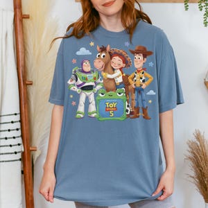May include: A blue t-shirt featuring characters from the animated film "Toy Story 5". The design includes Buzz Lightyear, Woody, Jessie, and a horse, with the movie title and number displayed on a green sign. The shirt is a comfortable, oversized fit.