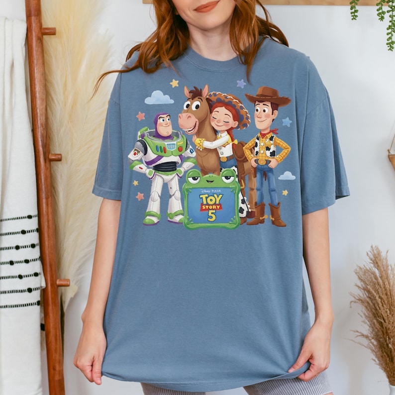 May include: A blue t-shirt featuring characters from the animated film "Toy Story 5". The design includes Buzz Lightyear, Woody, Jessie, and a horse, with the movie title and number displayed on a green sign. The shirt is a comfortable, oversized fit.