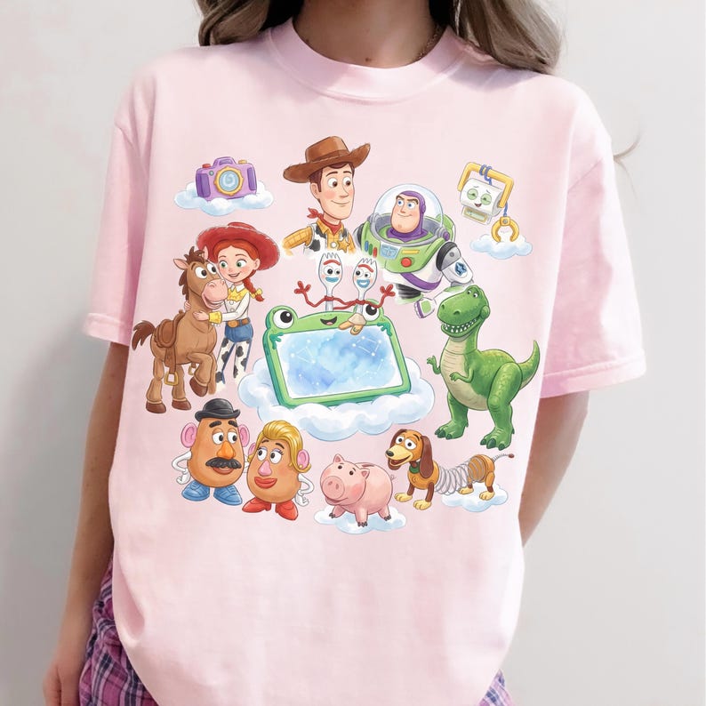 May include: Light pink t-shirt featuring a colorful graphic of characters from the animated movie Toy Story. The design includes Woody, Buzz Lightyear, Jessie, and other beloved characters, set against a cloud background.