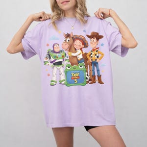 May include: A light purple t-shirt featuring a graphic of characters from the Toy Story franchise. The design includes Buzz Lightyear, Woody, Jessie, and a horse, with the text "Toy Story 5" on a green frog-shaped sign.