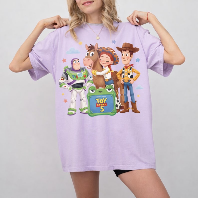 May include: A light purple t-shirt featuring a graphic of characters from the Toy Story franchise. The design includes Buzz Lightyear, Woody, Jessie, and a horse, with the text "Toy Story 5" on a green frog-shaped sign.