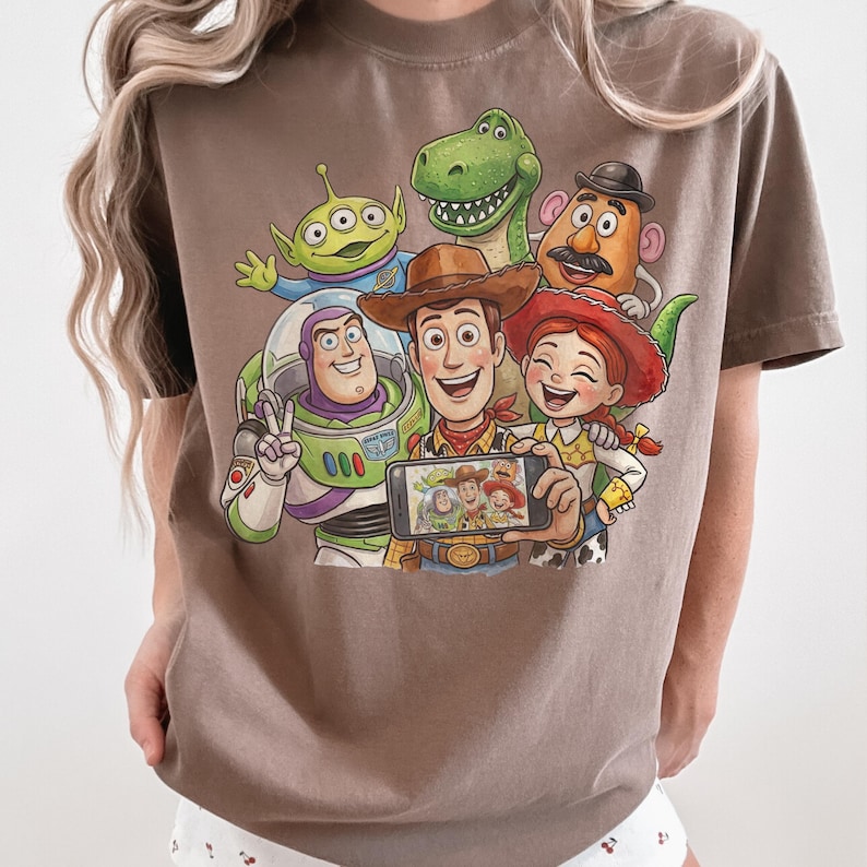 May include: A brown t-shirt featuring a graphic of characters from the animated film Toy Story. The design includes Woody, Buzz Lightyear, Jessie, Rex, Mr. Potato Head, and an alien, with Woody holding a phone displaying a selfie.