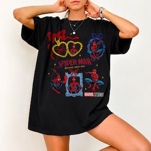 K&ouml;nnte beinhalten: Ein schwarzes T-Shirt mit einem Grafikdruck von Spider-Man in verschiedenen Posen, umrahmt von dekorativen R&auml;ndern und roten Schleifen. Der Text lautet Spider-Man Brand New Day und Marvel Studios, erg&auml;nzt durch kleine gelbe Sterne auf der Vorderseite.