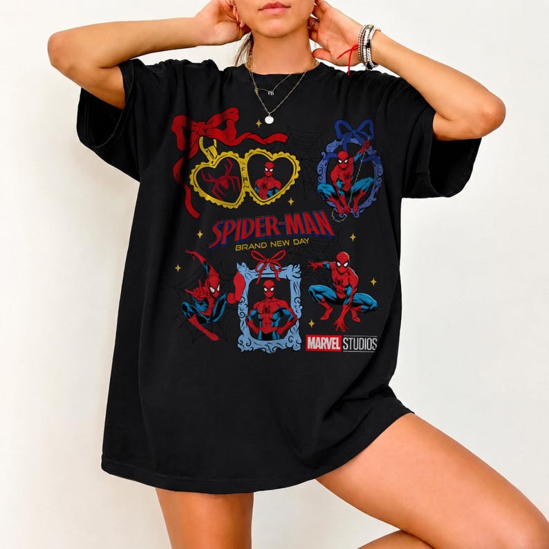 K&ouml;nnte beinhalten: Ein schwarzes T-Shirt mit einem Grafikdruck von Spider-Man in verschiedenen Posen, umrahmt von dekorativen R&auml;ndern und roten Schleifen. Der Text lautet Spider-Man Brand New Day und Marvel Studios, erg&auml;nzt durch kleine gelbe Sterne auf der Vorderseite.