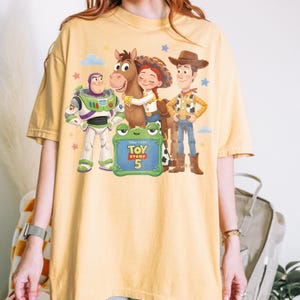 May include: Yellow t-shirt featuring a graphic of characters from the Toy Story movie franchise. The design includes Buzz Lightyear, Woody, Jessie, and a horse. A green sign reads "Toy Story 5".