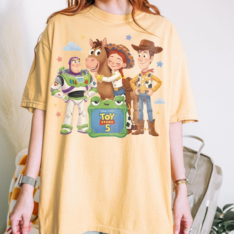 May include: Yellow t-shirt featuring a graphic of characters from the Toy Story movie franchise. The design includes Buzz Lightyear, Woody, Jessie, and a horse. A green sign reads "Toy Story 5".