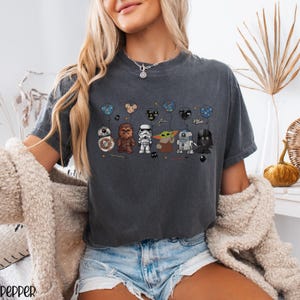 May include: Dark gray t-shirt featuring cartoon Star Wars characters holding balloon bouquets. Characters include BB-8, Chewbacca, a Stormtrooper, Grogu, R2-D2, and Darth Vader. The balloons have Mickey Mouse ears and other designs.