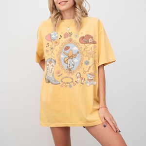 Puede incluir: Una camiseta de manga corta color amarillo mostaza con un estampado gr&aacute;fico central de una persona tocando la guitarra dentro de un marco ornamentado, rodeado de ilustraciones de una bota vaquera, un sombrero, un oso de peluche y flores.