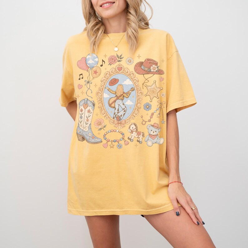 Puede incluir: Una camiseta de manga corta color amarillo mostaza con un estampado gr&aacute;fico central de una persona tocando la guitarra dentro de un marco ornamentado, rodeado de ilustraciones de una bota vaquera, un sombrero, un oso de peluche y flores.